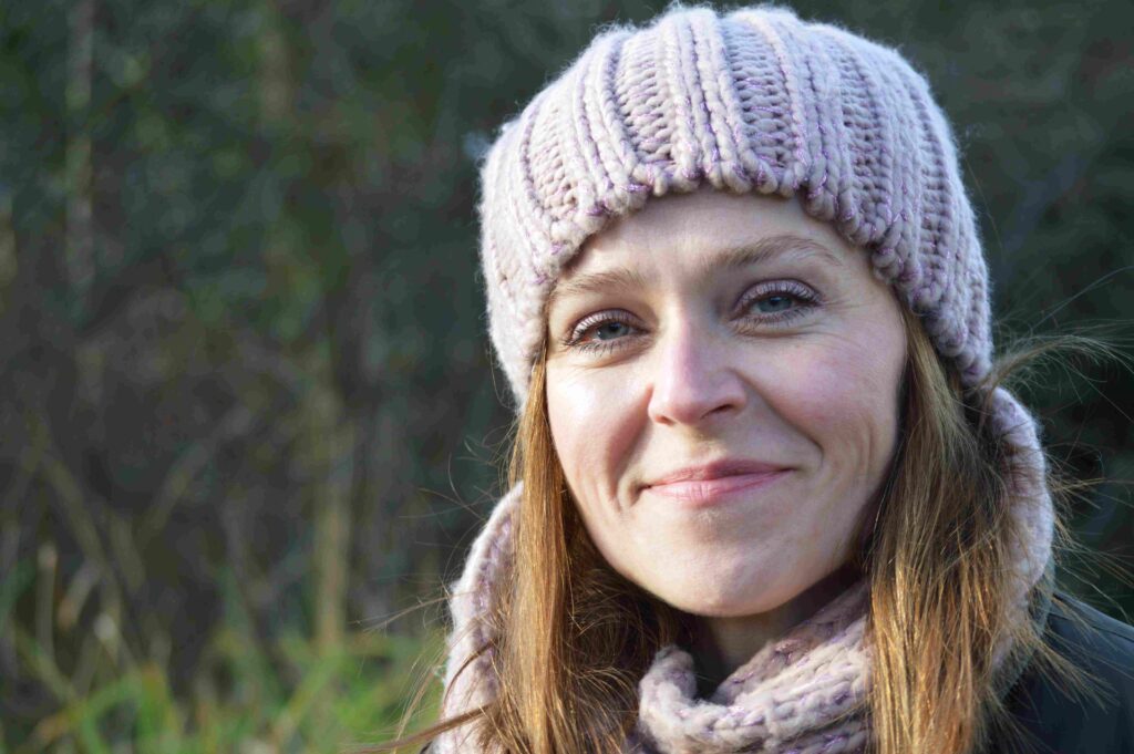 Close-up of a smiling woman wearing a chunky knit beanie and matching scarf outdoors on a cool day.