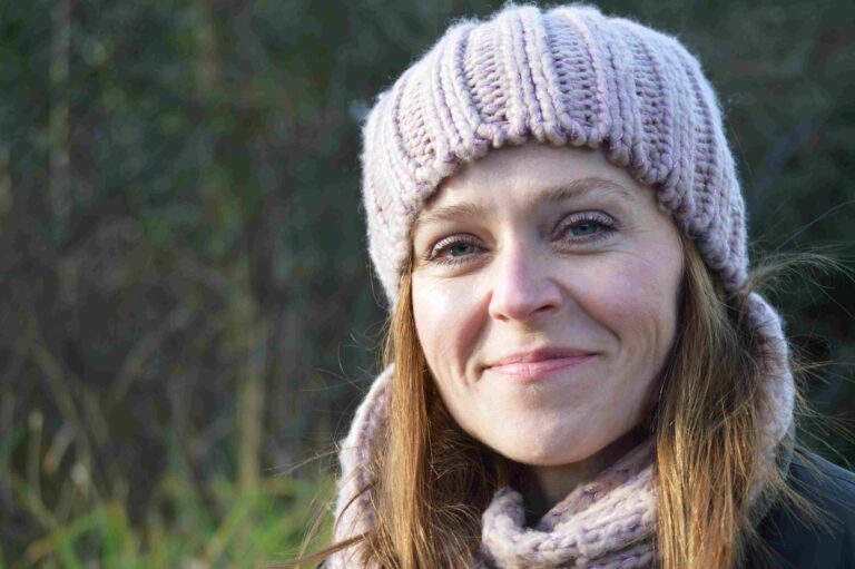 Close-up of a smiling woman wearing a chunky knit beanie and matching scarf outdoors on a cool day