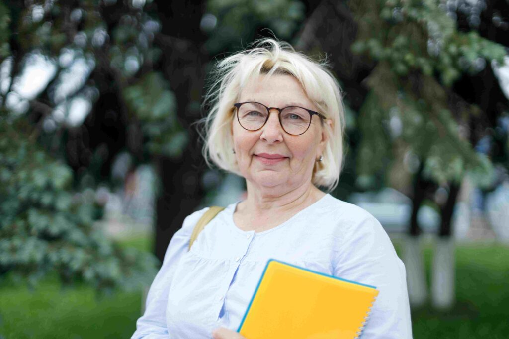 Smiling older woman with blond hair and glasses standing outside, holding a bright yellow notebook against a leafy background
