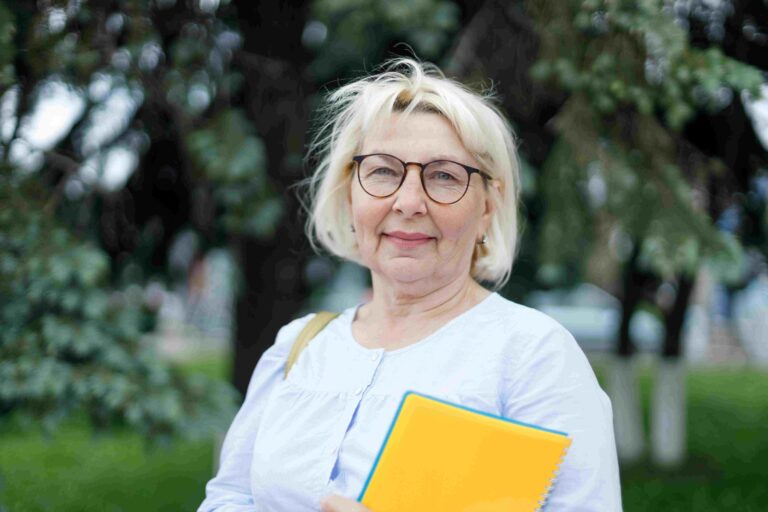 Smiling older woman with blond hair and glasses standing outside, holding a bright yellow notebook against a leafy background