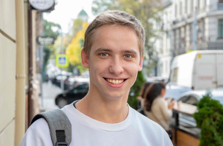 Smiling young man with a backpack strap over one shoulder standing on a city street.