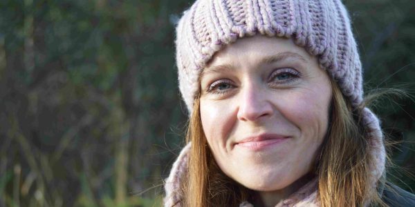 Close-up of a smiling woman wearing a chunky knit beanie and matching scarf outdoors on a cool day.