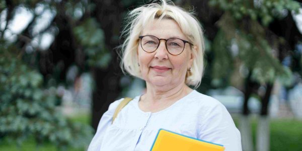Smiling older woman with blond hair and glasses standing outside, holding a bright yellow notebook against a leafy background