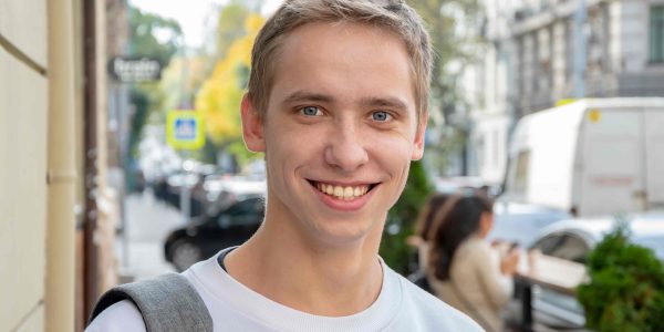 Smiling young man with a backpack strap over one shoulder standing on a city street.